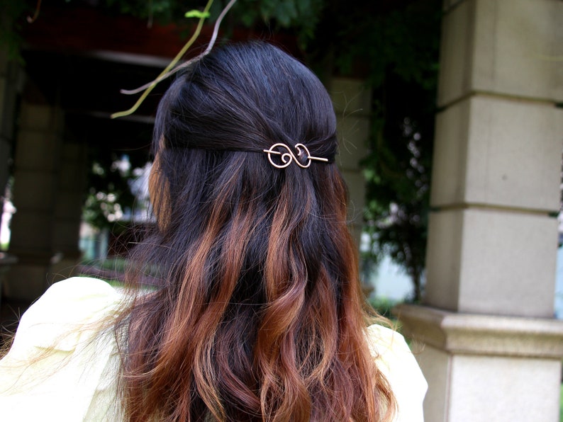 May include: A gold hair clip with a double loop design. The clip is in the hair of a person with long brown and black hair.