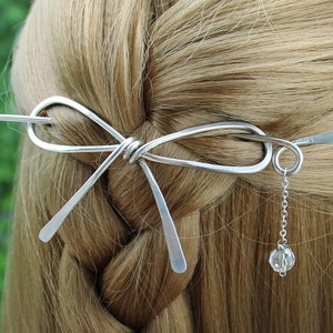 Op de afbeelding: Zilveren haarspeld in de vorm van een strik met een hangende kristallen hanger. De speld zit in het haar van een vrouw.