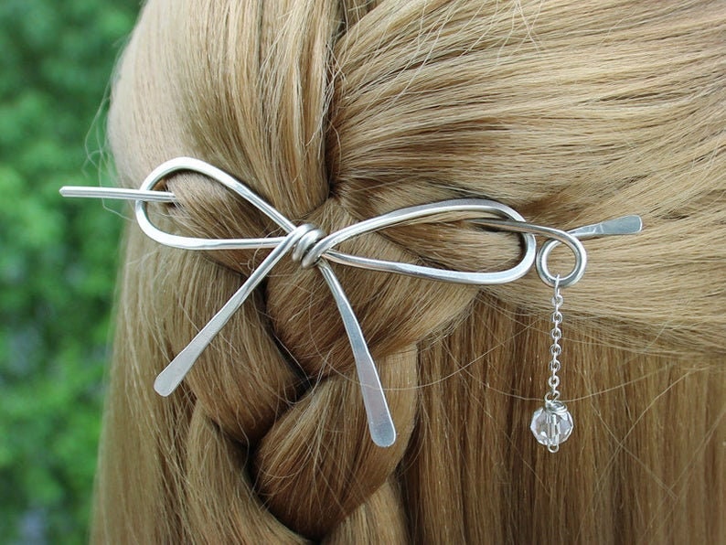 Op de afbeelding: Zilveren haarspeld in de vorm van een strik met een hangende kristallen hanger. De speld zit in het haar van een vrouw.