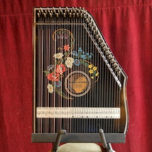 May include: An autoharp in black and gold, featuring strings, floral designs, and the word "Tone" within a circle. The instrument is set against a red backdrop. It is supported by a metal stand. The autoharp has a traditional design.