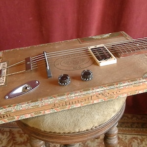 May include: A handmade cigar box guitar with a wooden body and a fretboard. The guitar features strings, pickups, and control knobs. The box has decorative floral patterns and the text "MAR.FIESTA" and "Sentiment Moor".