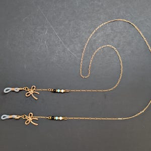 May include: Gold-coloured glasses chain with a delicate chain design. Each end has a bow-shaped charm and small beads in black, white, and turquoise. The chain is designed to hold spectacles.