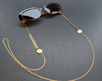 May include: Oval-shaped sunglasses with a tortoiseshell pattern, paired with a gold-colored chain. The chain features a leaf design and circular accents, creating a stylish accessory. The sunglasses rest on a dark surface.