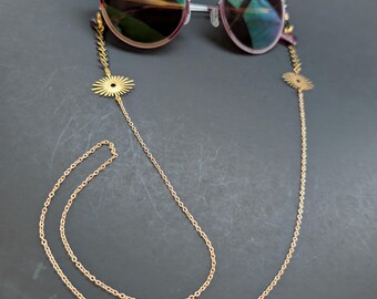 May include: Round-framed eyeglasses with a gold chain eyeglass holder. The chain features a delicate design with small, sun-shaped accents. The lenses have a gradient tint, and the frames are a mix of silver and pink.