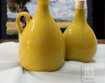 May include: Two yellow ceramic oil bottles with cork stoppers. The bottles have a rounded shape and a handle. The bottles are sitting on a white surface. The text "Arte Ceramica Pugliese" and "Terreccotte Realizzate a Mano" is visible in the image.