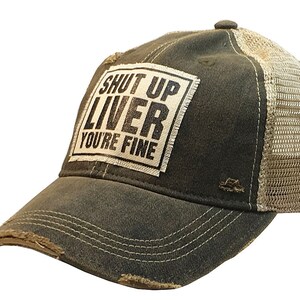 May include: A dark green and beige trucker hat with a distressed look. The front of the hat features a white patch with black text that reads "Shut Up Liver You're Fine".