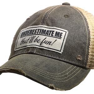 May include: A distressed black and brown trucker hat with a white patch that reads "UNDERESTIMATE ME that'll be fun!"