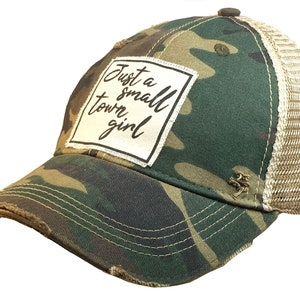 May include: A camouflage trucker hat with a distressed look. The hat has a white patch with the text "Just a small town girl" in black.