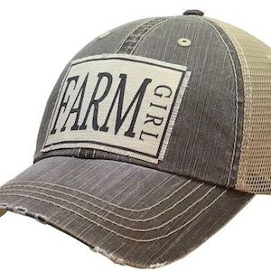 May include: A gray and beige trucker hat with a distressed look. The front of the hat has a white patch with black text that reads "FARM GIRL".