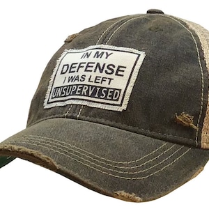 May include: Distressed brown baseball cap with a tan mesh back. The front patch reads "IN MY DEFENSE I WAS LEFT UNSUPERVISED" in black text. The brim and edges have a worn, frayed look.