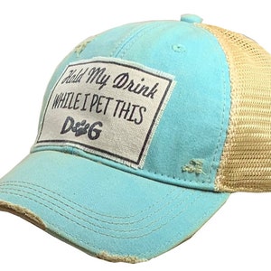 May include: A turquoise and beige trucker hat with a distressed look. The hat has a patch on the front that says "Hold My Drink While I Pet This Dog".