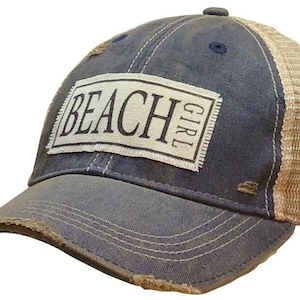 May include: Distressed blue baseball cap with a tan mesh back. The front features a white rectangular patch with the words "BEACH GIRL" in black. The brim and edges of the cap show signs of wear, giving it a vintage look.