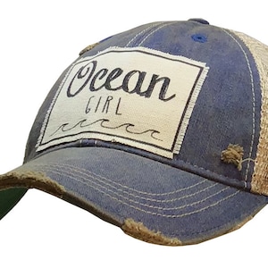 May include: A blue and beige distressed trucker hat with a white patch that says "Ocean Girl" with a wave design underneath.