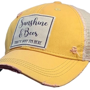 May include: Yellow trucker hat with a distressed look. The front panel features a rectangular patch with the words "Sunshine & Beer, That's Why I'm Here". The hat has a mesh back and a curved brim.