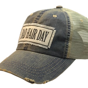 Op de afbeelding: Een blauwe denim en mesh trucker cap met een verweerde look. De pet heeft een witte patch met de tekst "Bad Hair Day" in zwart.