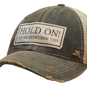 May include: Distressed olive green and tan trucker hat with the text "HOLD ON! LET ME OVERTHINK THIS" on a rectangular patch. The hat has a frayed brim and a mesh back.