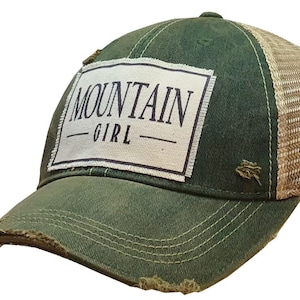 May include: A green and brown distressed trucker hat with a white patch that says "MOUNTAIN GIRL" in black lettering.