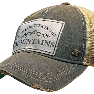 May include: A dark green and beige trucker hat with a distressed look. The hat has a patch on the front that says "Life is better in the mountains" with a mountain range graphic.