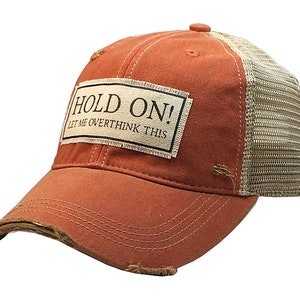 May include: A distressed orange trucker hat with a beige mesh back. The hat features a rectangular patch with the text "HOLD ON! LET ME OVERTHINK THIS". The brim has a distressed, worn look.