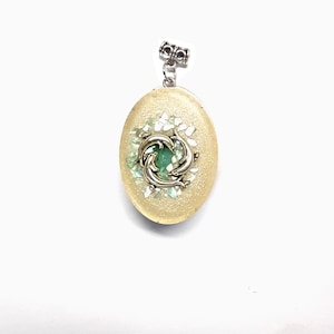 May include: A light yellow oval pendant with a silver dolphin charm and a green stone in the center. The pendant is surrounded by white glitter.
