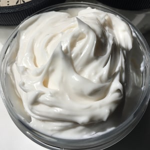 May include: Close-up of a white, creamy body butter in a clear glass jar. The butter is thick and smooth, with a swirl pattern.