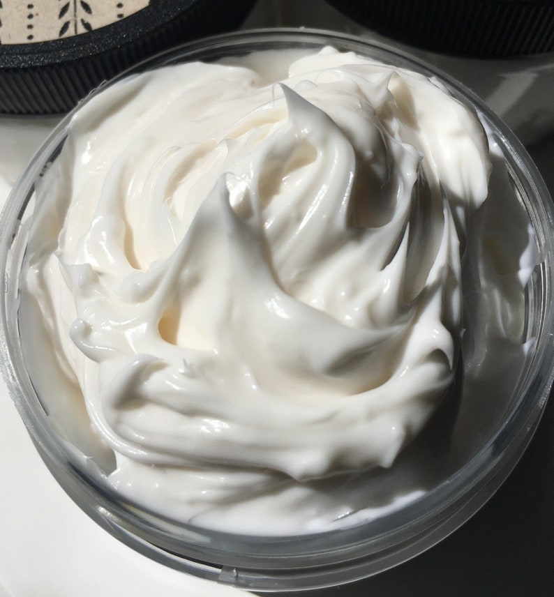 May include: Close-up of a white, creamy body butter in a clear glass jar. The butter is thick and smooth, with a swirl pattern.