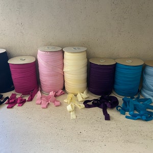 May include: Eight spools of elastic ribbon in various colors, including black, pink, light pink, cream, purple, blue, and light blue. The spools are arranged in a row on a light-colored surface.