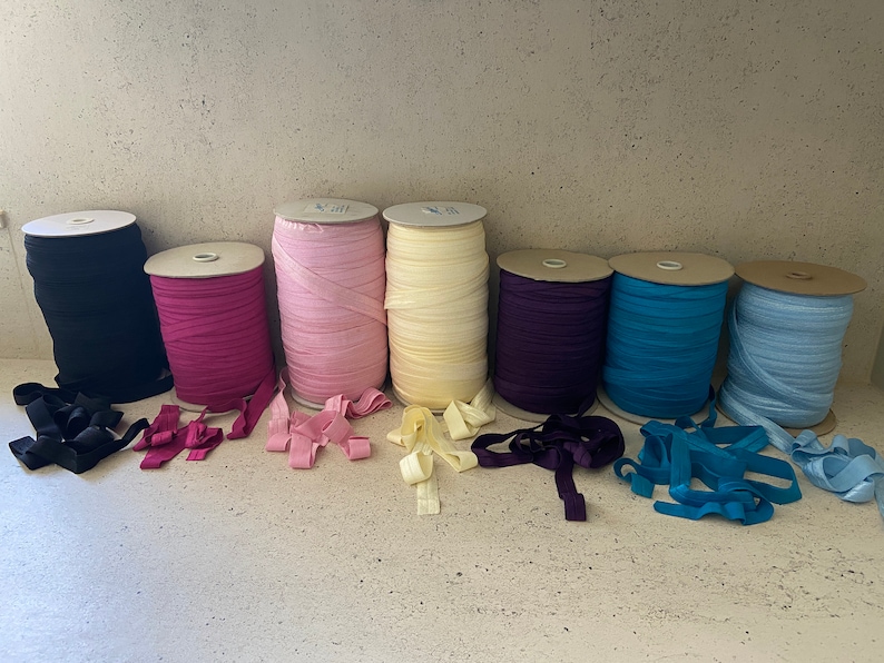 May include: Eight spools of elastic ribbon in various colors, including black, pink, light pink, cream, purple, blue, and light blue. The spools are arranged in a row on a light-colored surface.