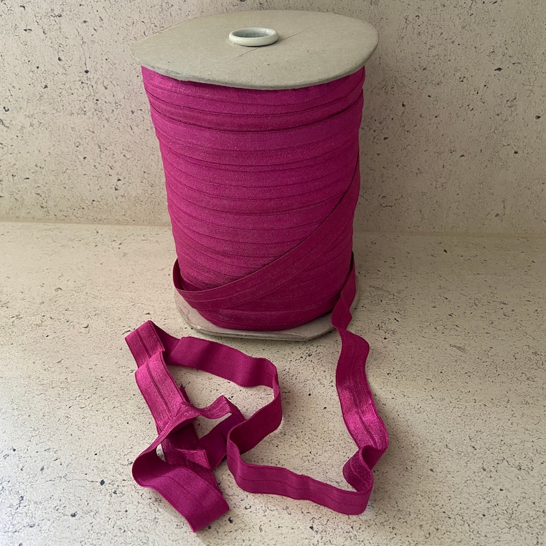 May include: A spool of pink elastic fabric, wound around a cardboard tube. The elastic is folded and laid out in a loose loop.