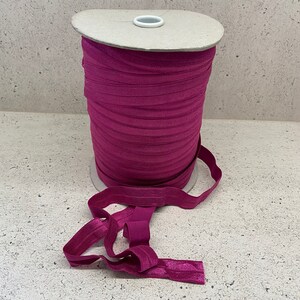 May include: A spool of magenta elastic band, wound around a cardboard core. The elastic is folded and laid out on a light gray surface.
