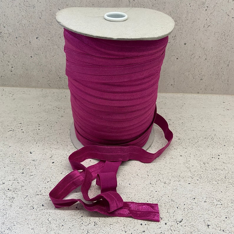 May include: A spool of magenta elastic band, wound around a cardboard core. The elastic is folded and laid out on a light gray surface.