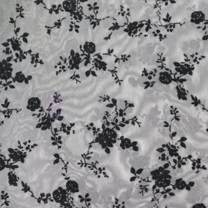 Könnte beinhalten: Nahaufnahme eines weißen Stoffes mit einem schwarzen Blumenmuster. Das Muster zeigt Rosen und andere Blumen mit Blättern und Stielen. Der Stoff hat ein leicht strukturiertes Aussehen.