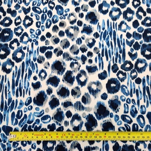 May include: A fabric with a blue and white leopard print design. The pattern features various shades of blue spots and brushstrokes against a white background. A yellow measuring tape is at the bottom of the image, showing inches.