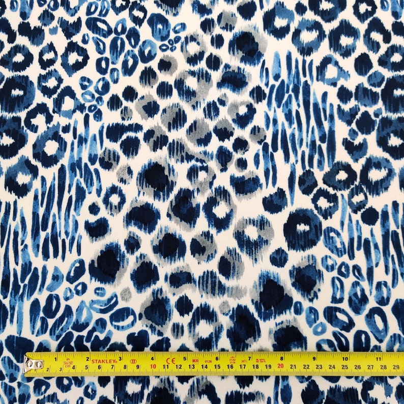 May include: A fabric with a blue and white leopard print design. The pattern features various shades of blue spots and brushstrokes against a white background. A yellow measuring tape is at the bottom of the image, showing inches.