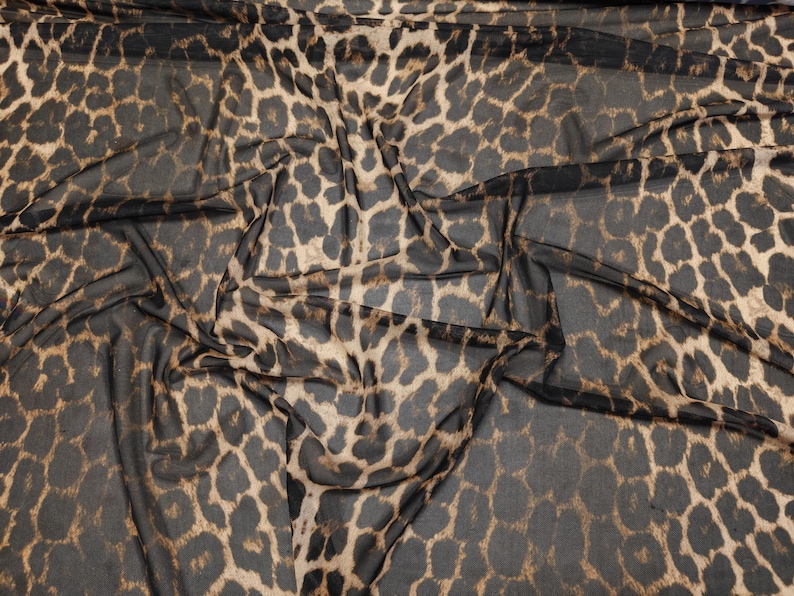 May include: Close-up of a sheer fabric with a leopard print pattern. The fabric features a dark brown and tan spotted design on a black background. The material is draped, showing the texture and flow of the fabric.
