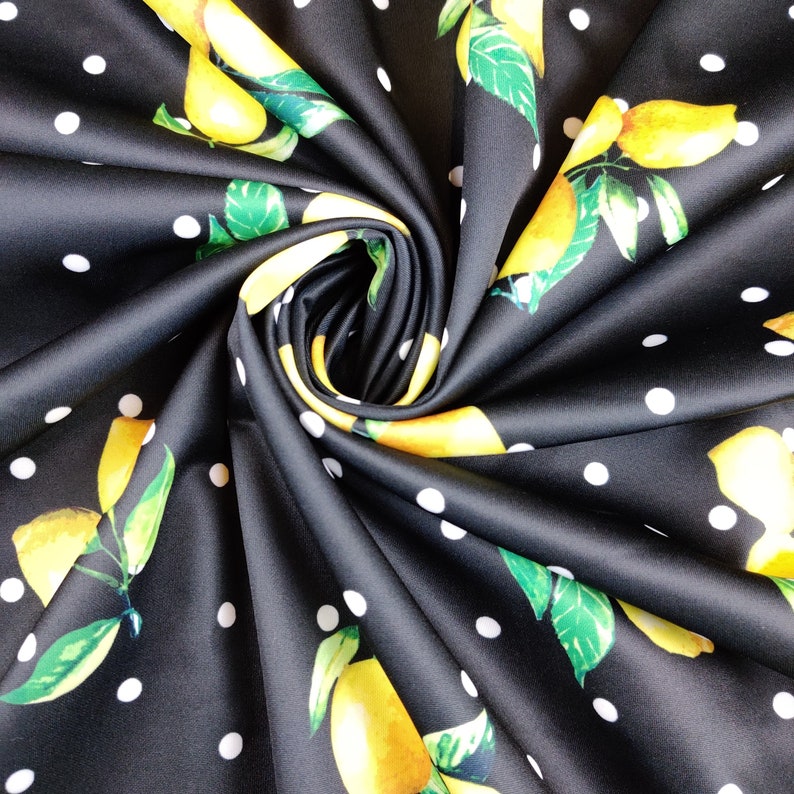 May include: A black fabric featuring a repeating pattern of bright yellow lemons with green leaves and small white polka dots. The material is gathered into a central spiral, showcasing the vibrant fruit print and the contrasting white spots.