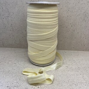 May include: A roll of pale yellow elastic, also known as fold over elastic, is wound around a cardboard core. The elastic is a soft, stretchy fabric with a subtle sheen.