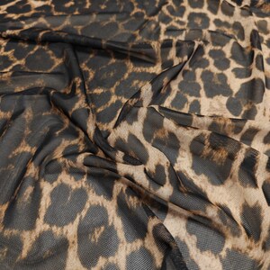 May include: Close-up of a sheer fabric with a leopard print pattern. The fabric is a mix of black and tan, with the leopard spots varying in size and shape. The material is draped, creating shadows and depth.