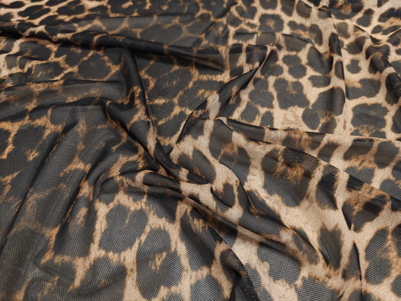 May include: Close-up of a sheer fabric with a leopard print pattern. The fabric is a mix of black and tan, with the leopard spots varying in size and shape. The material is draped, creating shadows and depth.