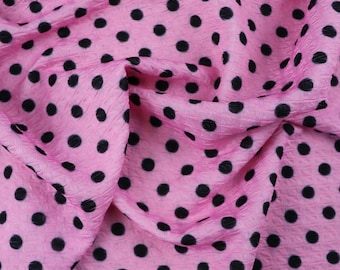 Peut inclure: Un tissu texturé présentant un motif répétitif de pois noirs unis sur un fond rose vif. Le matériau présente une finition de surface distincte, plissée ou gaufrée, avec des plis visibles et un drapé souple sur l'ensemble de la pièce.