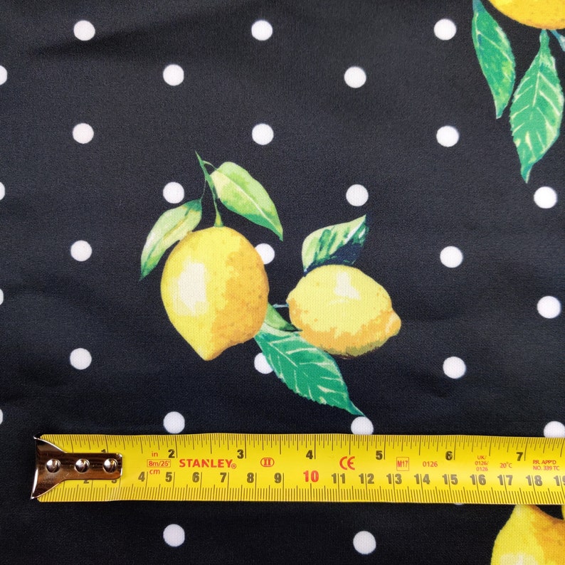 May include: A black fabric featuring a white polka dot pattern and a yellow lemon print with green leaves. A yellow Stanley measuring tape is positioned at the bottom, showing a length of approximately 7.5 inches across the fabric surface.