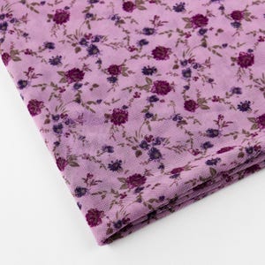 May include: A folded fabric piece featuring a detailed floral design. The fabric is a pale lavender, patterned with clusters of dark purple and blue flowers, and green leaves. The pattern is repeated across the material, creating a delicate aesthetic.