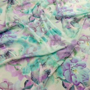 May include: Close-up of a fabric with a floral pattern. The design features large, stylized flowers in shades of purple, teal, and white, creating a watercolour effect. The fabric is draped, showing the texture and flow of the material.