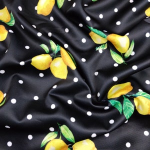 May include: A black fabric featuring a repeating pattern of yellow lemons with green leaves and small white polka dots. The material has a smooth texture with visible folds and drapes that create shadows and highlights across the surface.