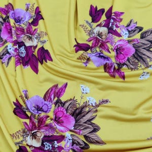 May include: Yellow fabric with a floral pattern. The design features clusters of pink, purple, and white flowers with dark purple and brown leaves. The fabric has a smooth, slightly shiny texture.