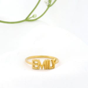 Puede incluir: Una taza de cerámica blanca con asa negra y el texto "But First, Coffee" en negrita negra. La taza se encuentra sobre un fondo borroso, sugiriendo un ambiente acogedor.