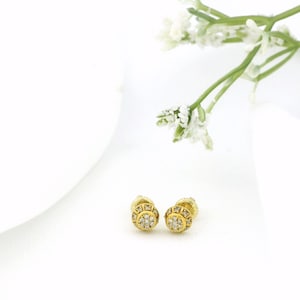 May include: A pair of gold-colored stud earrings with a circular design. Each earring features a central cluster of small, clear stones surrounded by a ring of smaller stones. The earrings are set against a white background with a sprig of white flowers.