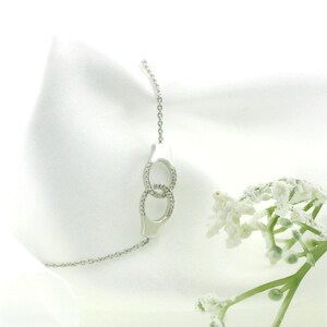 Puede incluir: Un collar plateado con un colgante formado por dos círculos entrelazados, cada uno incrustado con pequeñas piedras preciosas transparentes. El collar se muestra sobre un fondo de tela blanca, con flores blancas en primer plano.
