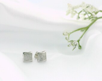 May include: A pair of square silver stud earrings with a grid of small, clear stones. The earrings are set against a white background with a sprig of white flowers.