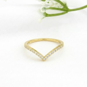 May include: A gold V-shaped ring, set with small, clear gemstones along the top edge. The ring is displayed on a white background, with a sprig of white flowers in the upper right corner.